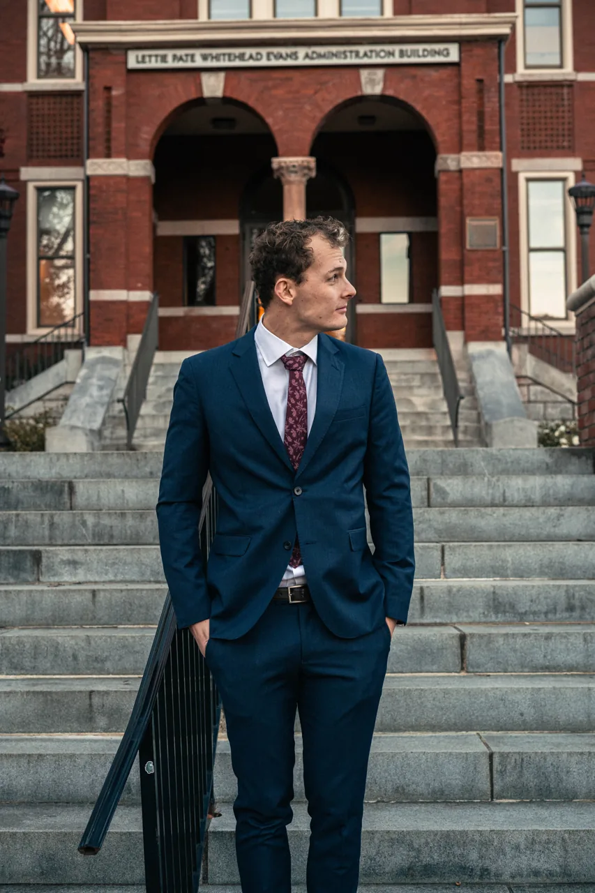 Daniel wearing a suit and graduation stole outside a university building.